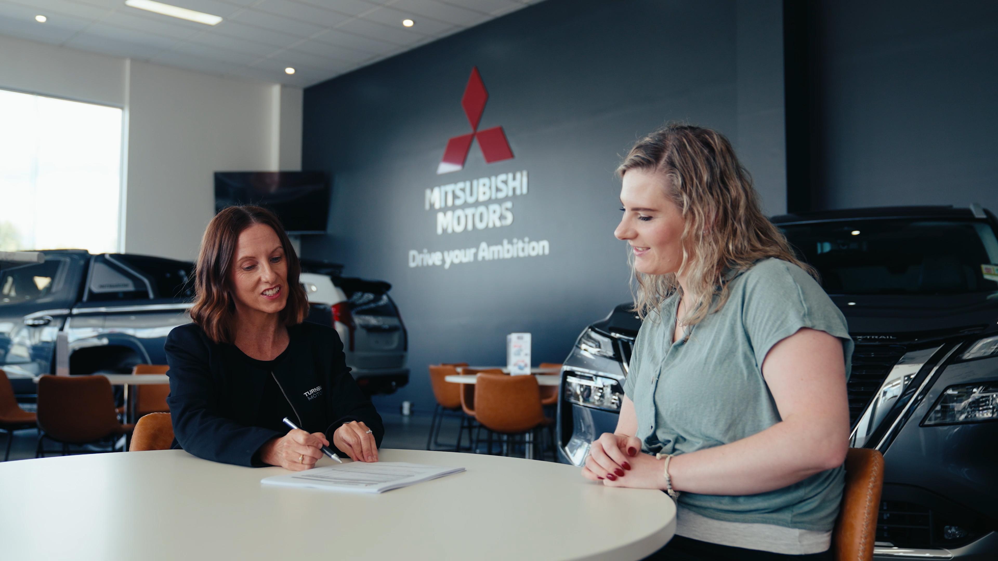 Una persona revisando una cotización de financiamiento automotriz en una agencia. En la pantalla o documento deben aparecer conceptos como enganche, mensualidad, plazo, tasa y CAT. La escena puede mostrar una agencia Mitsubishi de fondo, pero no es indispensable incluir un vehículo.
